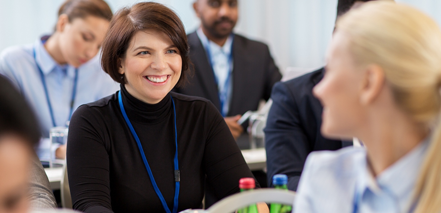 Woman sitting down at a fonference turning around talking to another woman.