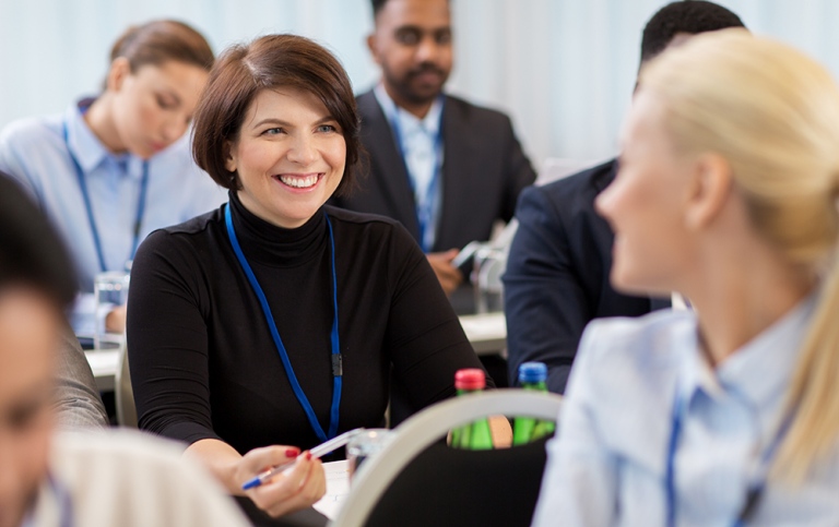 Woman sitting down at a fonference turning around talking to another woman.