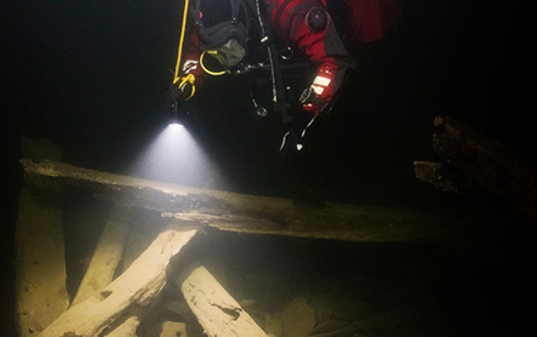 Under vatten. En dykare tittar neråt och lyser med en ficklampa på vrakdelar i trä. 