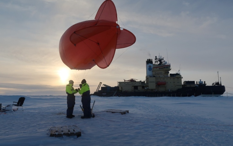 Scientists standing on the ice, handling a research balloon. An ice breaker in the background.