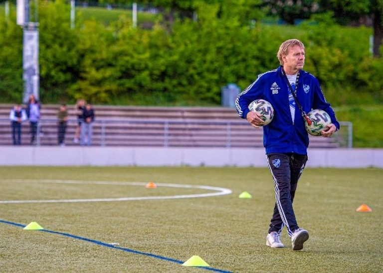 Man går på fotbollsplan bärande två fotbollar.
