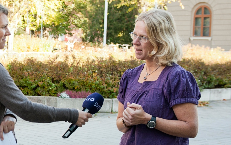 A journalist holds a microphone marked SVT in front of a researcher, a large TV camera stands next t