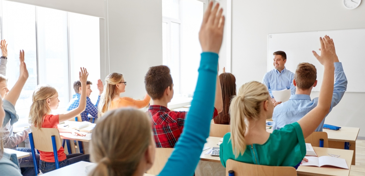 Students in a classroom all raising their hands, a male teacher smiling