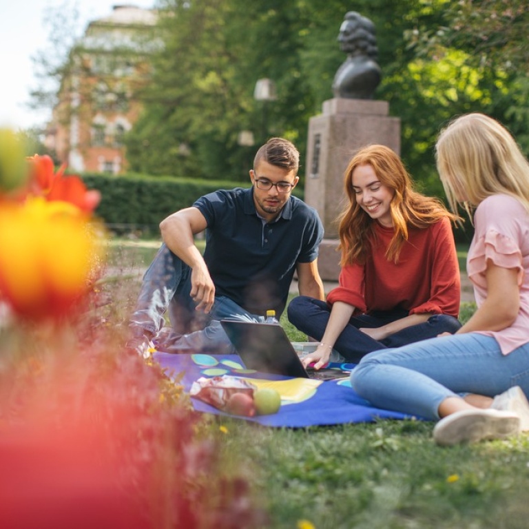 Tre studenter sitter på en filt på en gräsmatta och tittar i en laptop.