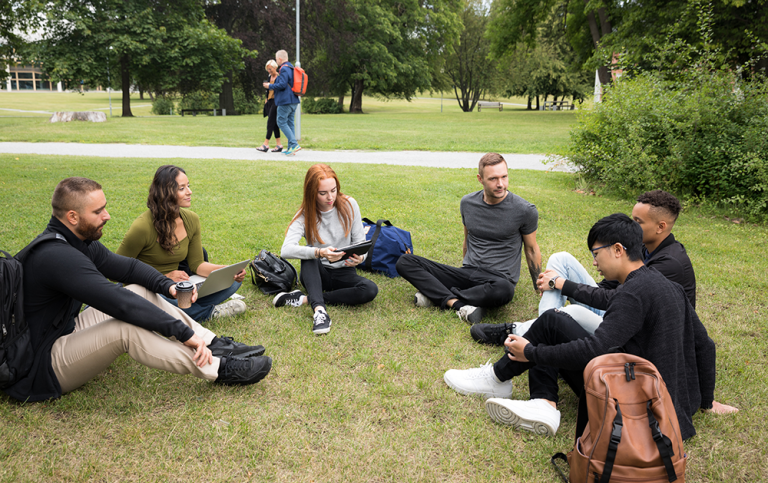 Sex studenter sitter i en ring på en gräsmatta på universitetsområdet.