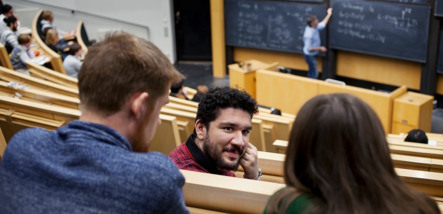 Conversation in auditorium, someone writing on blackboard in the background.