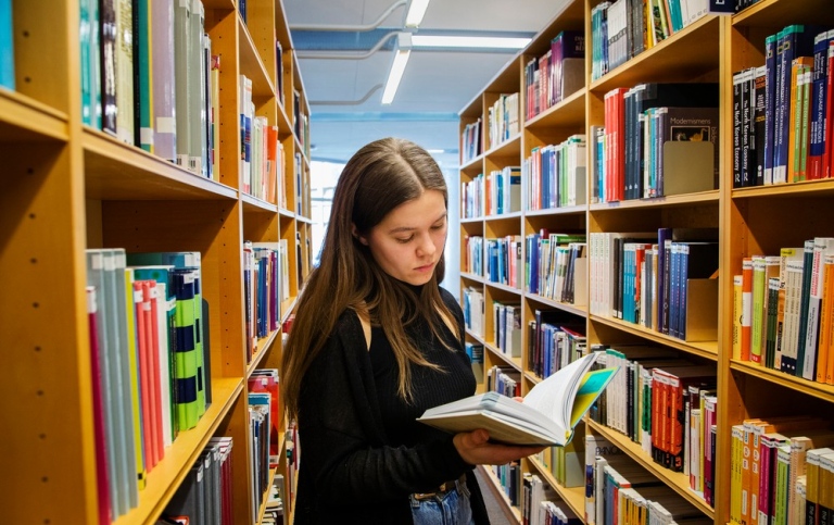 Kvinna står och läser i en bok mellan två bokhyllor på ett bibliotek