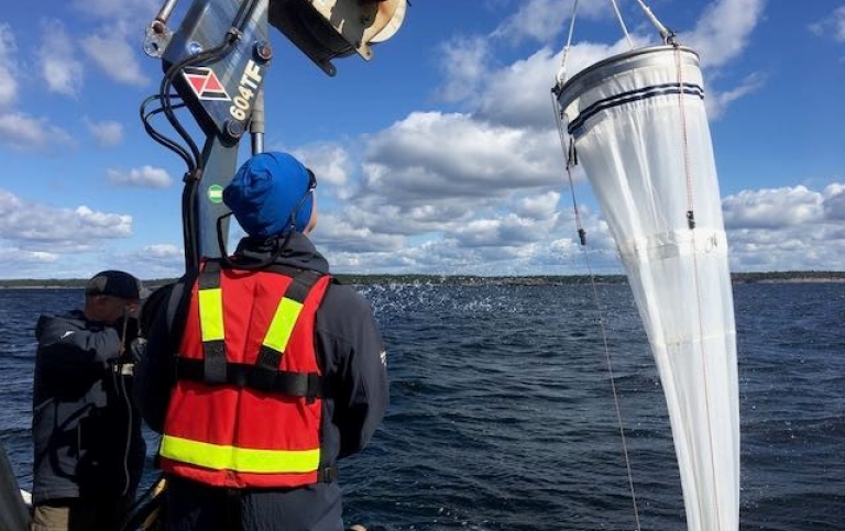 Man in a life jacket and blue hat on a boat is lifting a large filter with a crane.