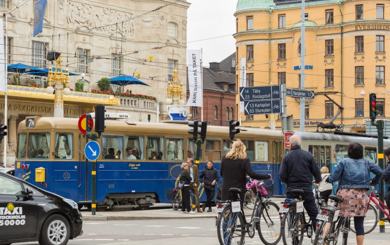 Genre photo of traffic in Stockholm with humans, bikes, cars and trams.