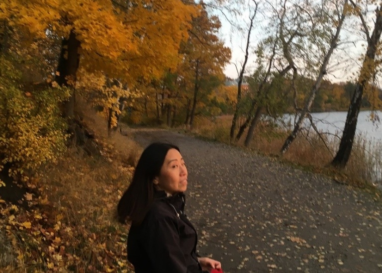 Woman with long dark hair and a black jacket walks along a foot path surrounded by trees in autumn