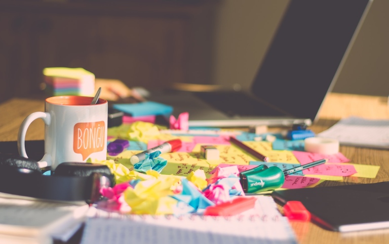 Oordnat skrivbord med dator, kaffekopp och post-it-lappar. Foto: Ferenc Horvath på Unsplash.