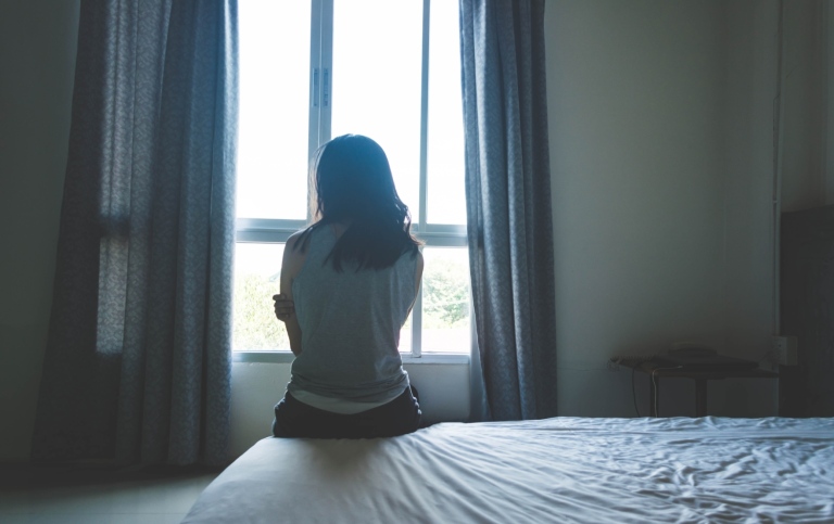 En kvinna sitter på en säng i ett rum vid ett fönster, med ryggen mot kameran. 
