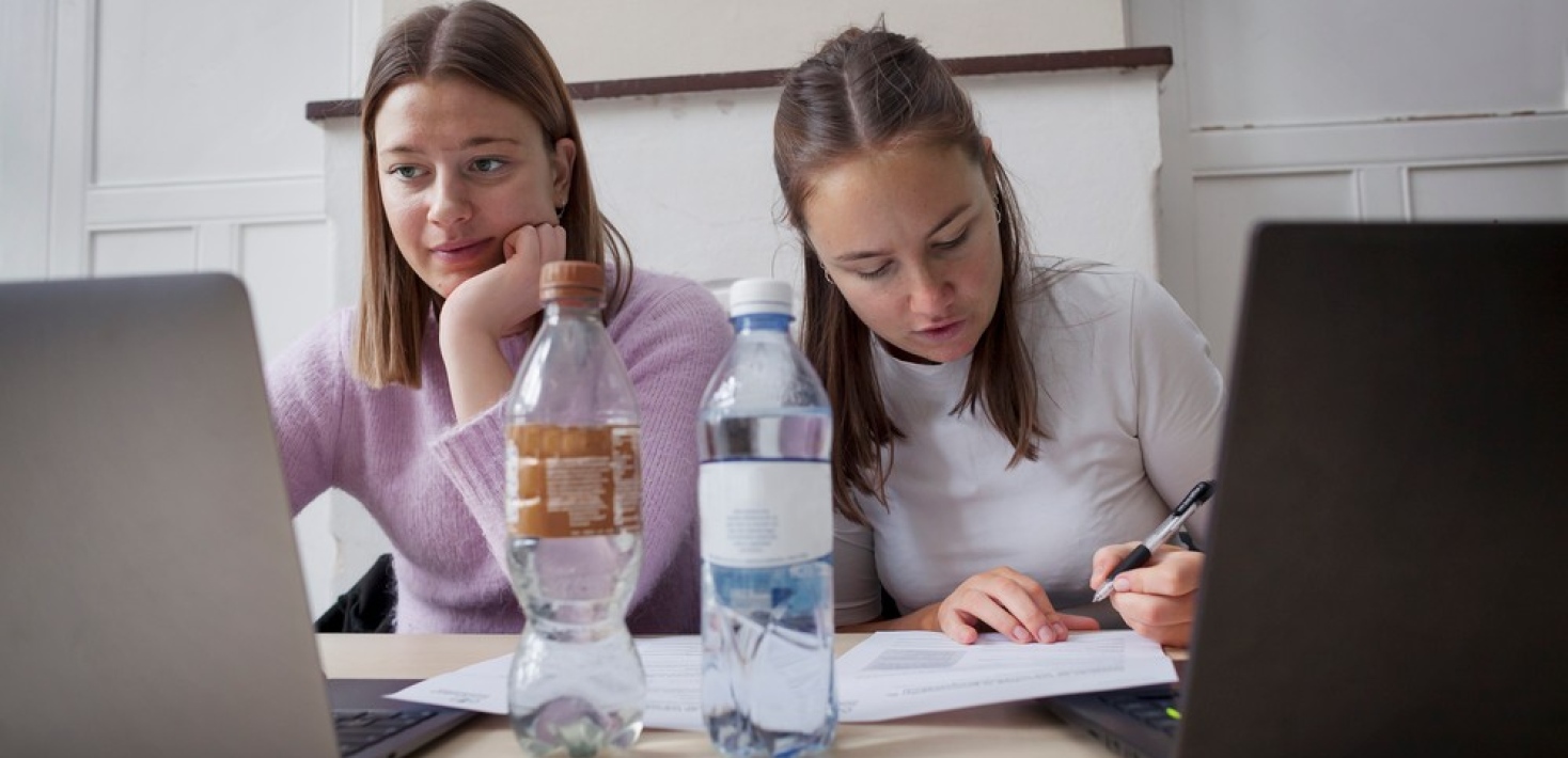 Två kvinnliga studenter sitter och pluggar bredvid varandra med laptops och vattenflaskor på bordet.