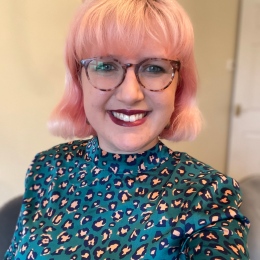 Ellie, a white woman wearing glasses with pink hair, smiles wearing a green leopard print dress. 