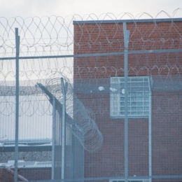 Prison fences, in the background a brick building and dull weather.