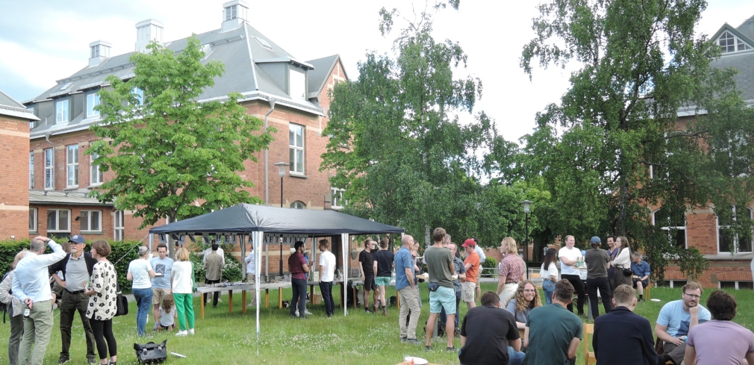 Sommarfest utomhus, folk som sitter på bänk och andra som står i olika grupper på gräsmatta.