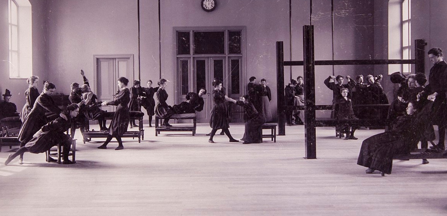 Svartvitt foto. Kvinnor i gymnastikhall gör gymnastiska övningar i gammaldags mörka kläder.