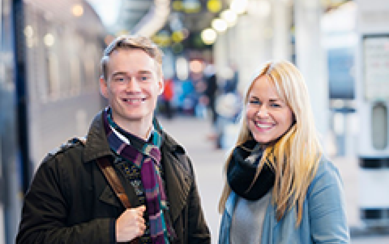 Två studenter står med väskor intill ett tåg på en perrong. De ler.