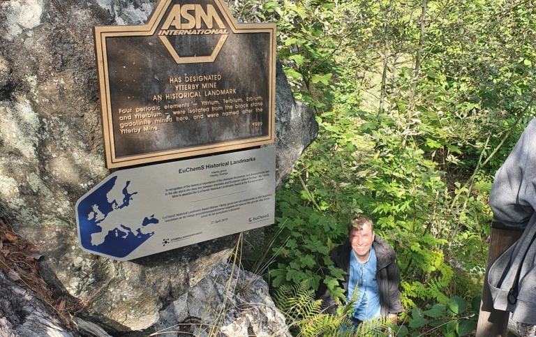 Teacher stands in a lot of bushes and trees below a plaque from ASM International