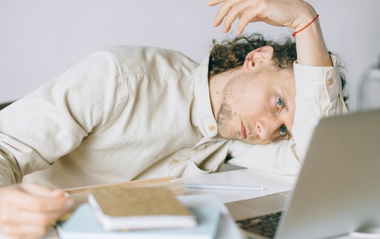 Tired man with pile of books and a computer. Photo: Nataliya Vaitkevich from Pexels