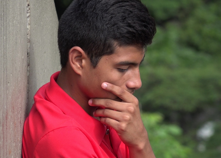 A dark-haired boy in red shirt sitting down, looking sad, with his left hand on his right cheek.