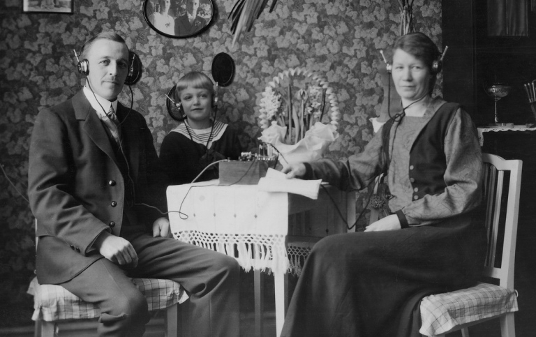 Svartvitt foto av två vuxna och ett barn med hörlurar och radioapparat av äldre modell