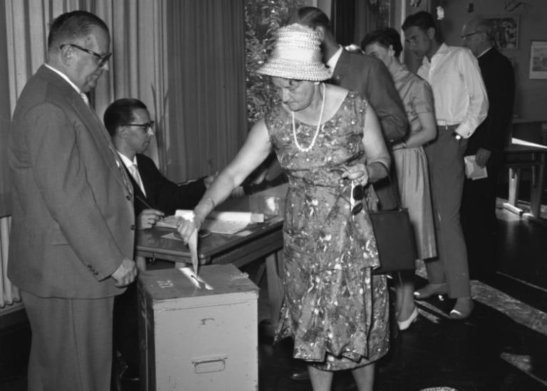 Svartvitt foto av kvinna i hatt som lägger en valsedel i en valurna