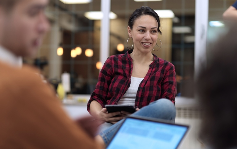 A woman in focus holding a tablet and smiling, a couple of other blurred people in front of her.