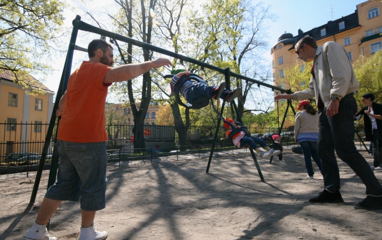 Två föräldralediga pappor gungar sina barn i lekparken på Nytorget i Stockholm. 