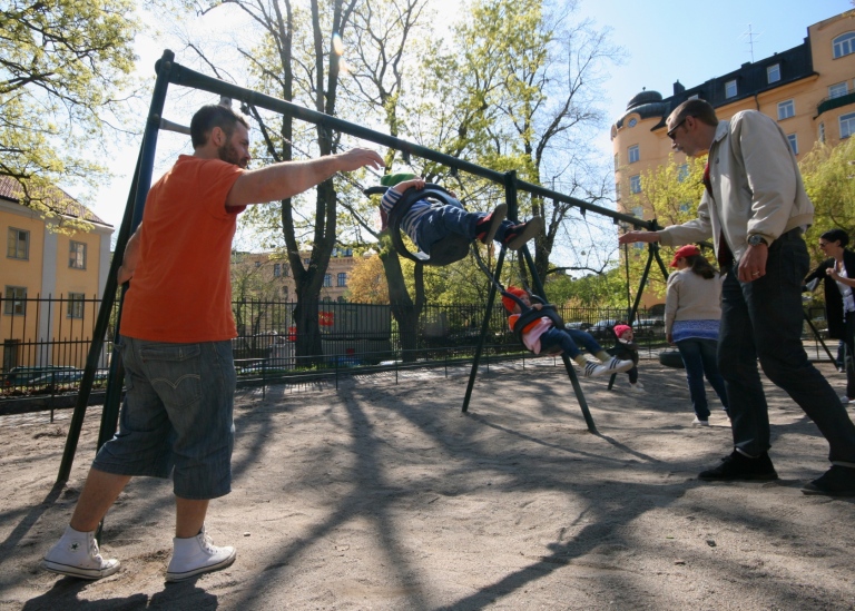 Two fathers on parental leave play with their children on the swings in a playground in Stockholm.