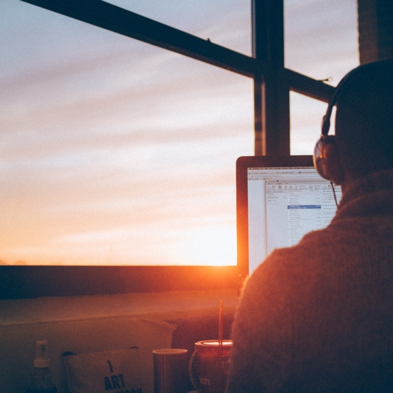 A man sitting in front of a computer from behind, wearing headphones, with a window in front of him.