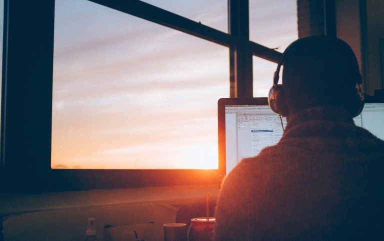 A man sitting in front of a computer from behind, wearing headphones, with a window in front of him.