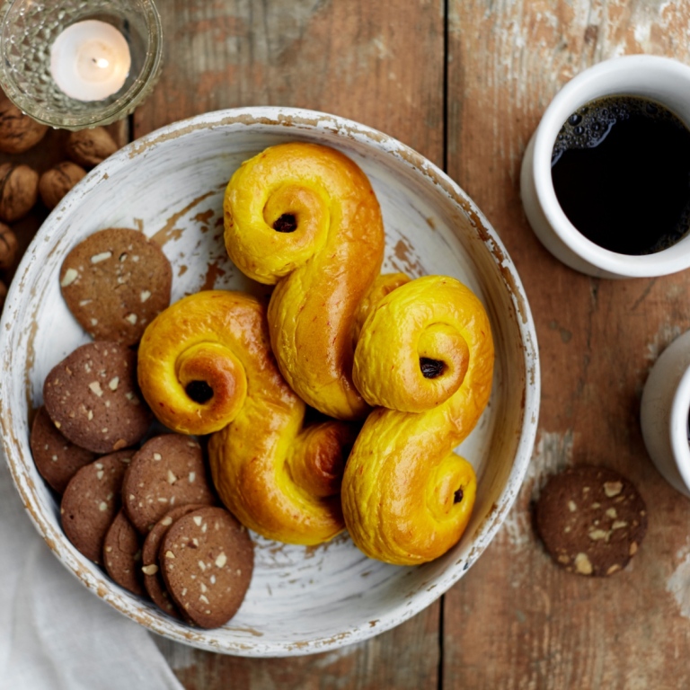 A place with saffron buns and cookies, two cups of coffee and other objects on a wooden table.
