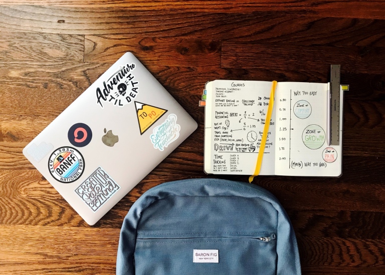 A blue backpack, a closed laptop with stickers, an open notebook with notes and a ruler on a desk.