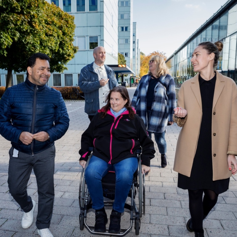 Två män och tre kvinnor pratar på campus. Kvinnan i mitten sitter i rullstol.