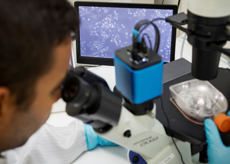 Forskare tittar i mikroskop med blå datorskärm i bakgrunden