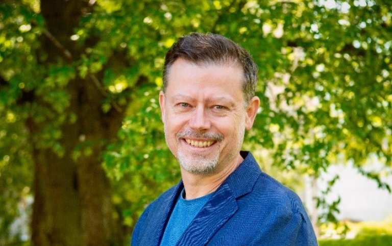 Man i Blå jacka framför grönskande träd.