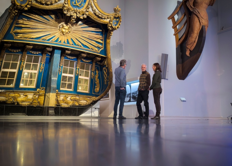 Tre personer står i en stor hall framför en vackert snidad akter på ett skepp.