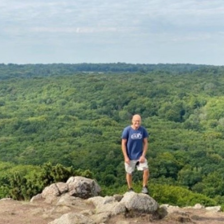 A man in a t-shirt and shorts stands on a rock on top of a hill, with a green view.