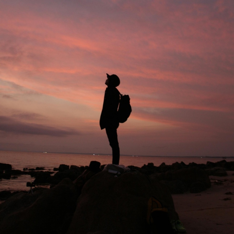 Siluett av ung kille som står på klippor vid havet i solnedgång.