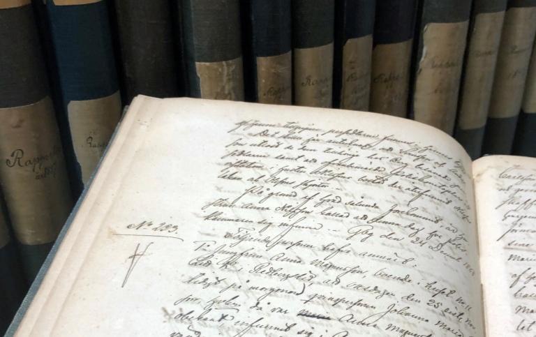 An open book with old, handwritten text in Swedish in front of a bookshelf.