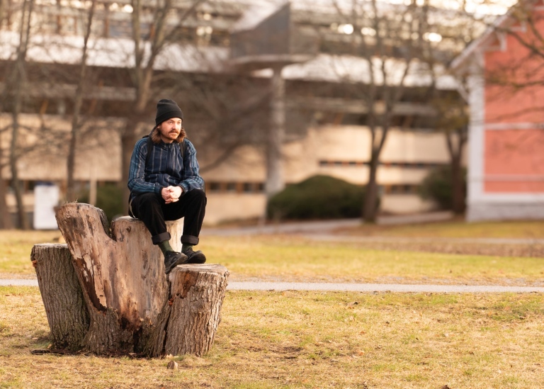Noa Persson sitter på en stubbe utanför Stockholms universitets byggnader.