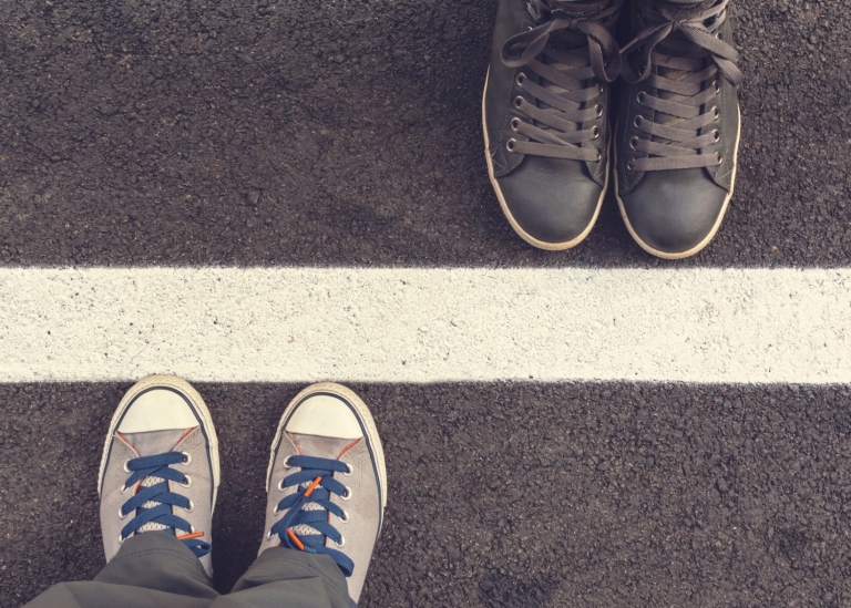 Bild på två par fötter, med avstånd från varandra, med en målad bred linje på asfalten mellan dem.
