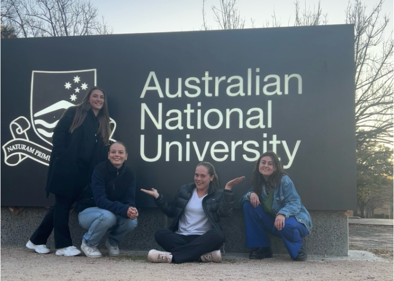 Foto på fyra tjejer framför en stor skylt med Australian National Universitys emblem och namn.