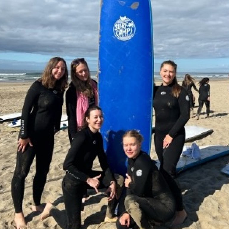 Bild på fem tjejer vid en surfbräda på en strand med havet i bakgrunden.