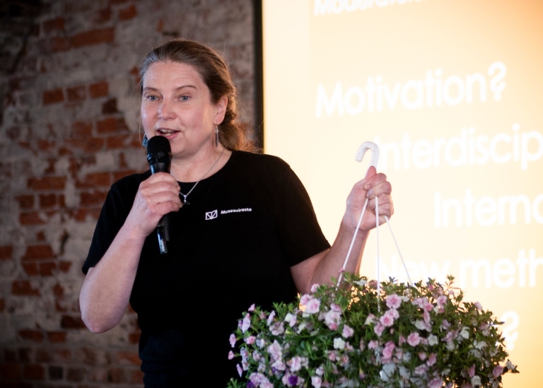 Minna Koivikko speaking into a microphone and holding a potted flower.