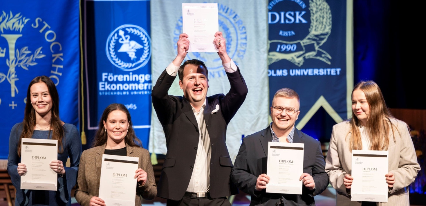 Fem glada studenter står på rad och visar upp sina diplom.
