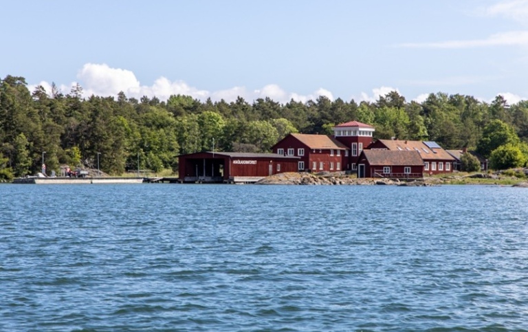 En samling rödmålade byggnader på en skärgårdsö, sedda från en båt.