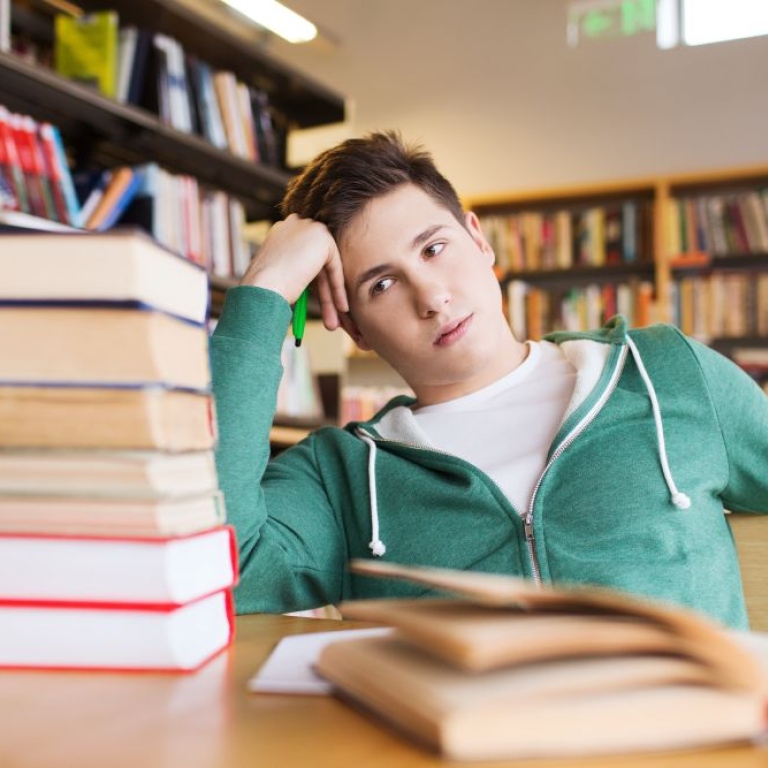 Male student looking at a pile of books, looking tired.