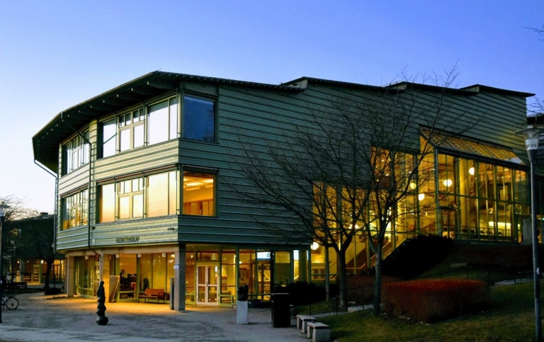 A green building with large windows glowing at dusk, surrounded by trees and a lit interior staircas